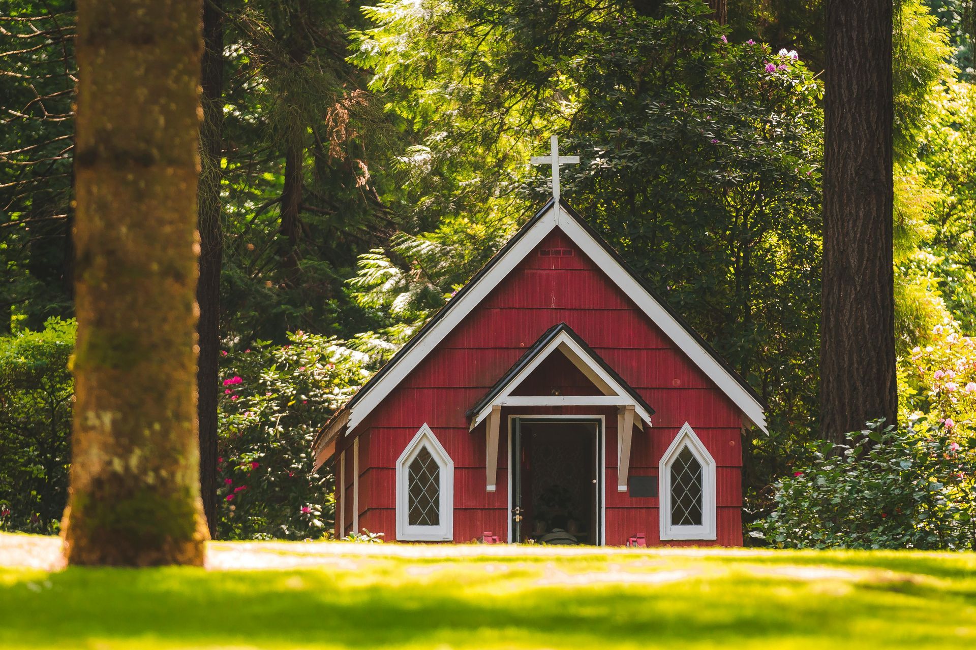 Red chapel with cross on the roof, surrounded by green trees and grass.
