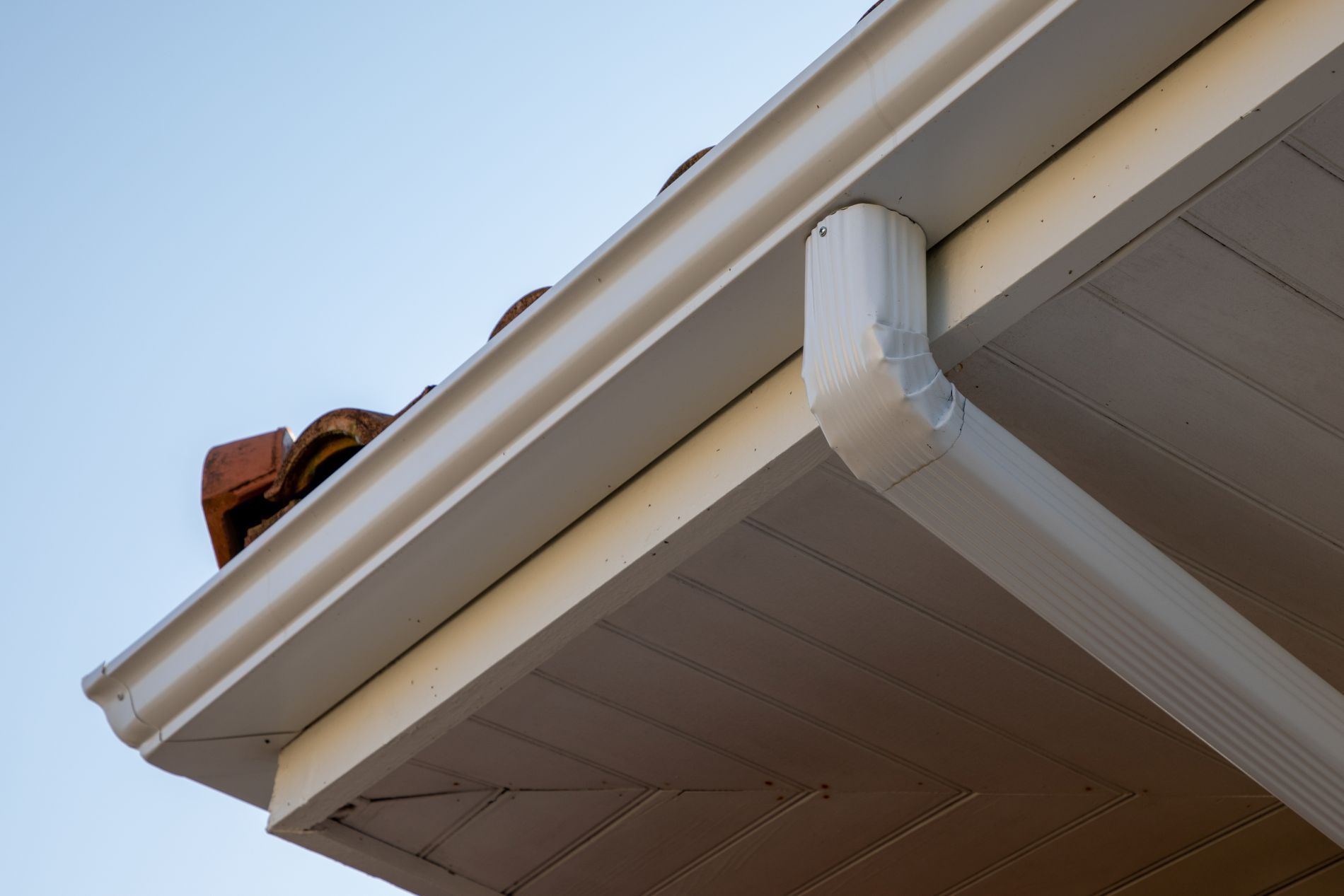 White gutter system attached to the roof of a house, angled down, against a blue sky.