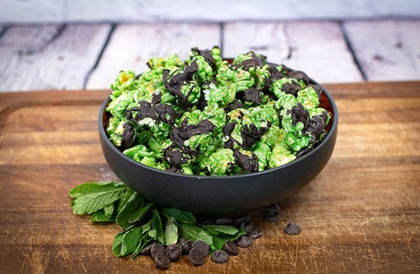 A bowl of green popcorn with chocolate chips and mint leaves on a wooden table.
