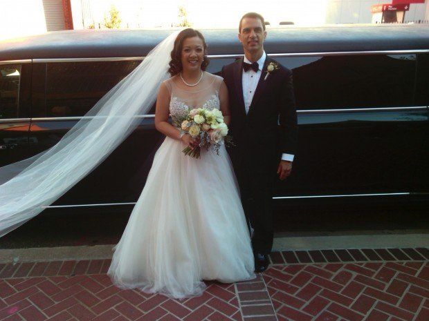 A bride and groom pose in front of a black limousine
