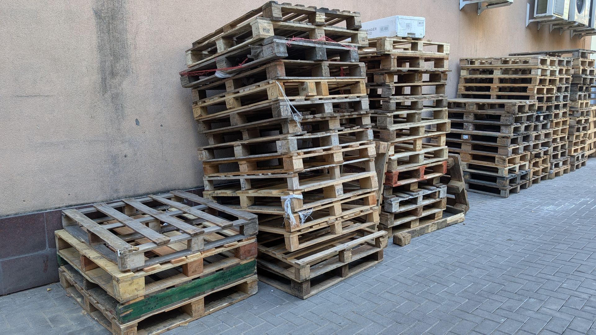Multiple stacks of worn wooden shipping pallets leaning against a beige wall on a paved surface.