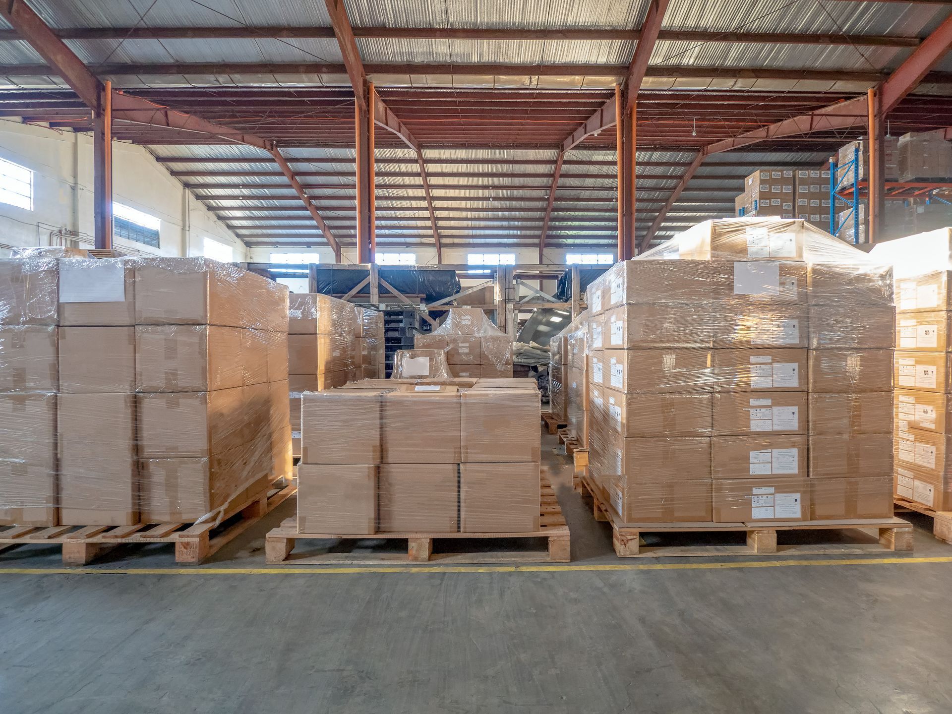 Three pallets of cardboard boxes shrink-wrapped for storage inside a large, industrial warehouse with metal beams.