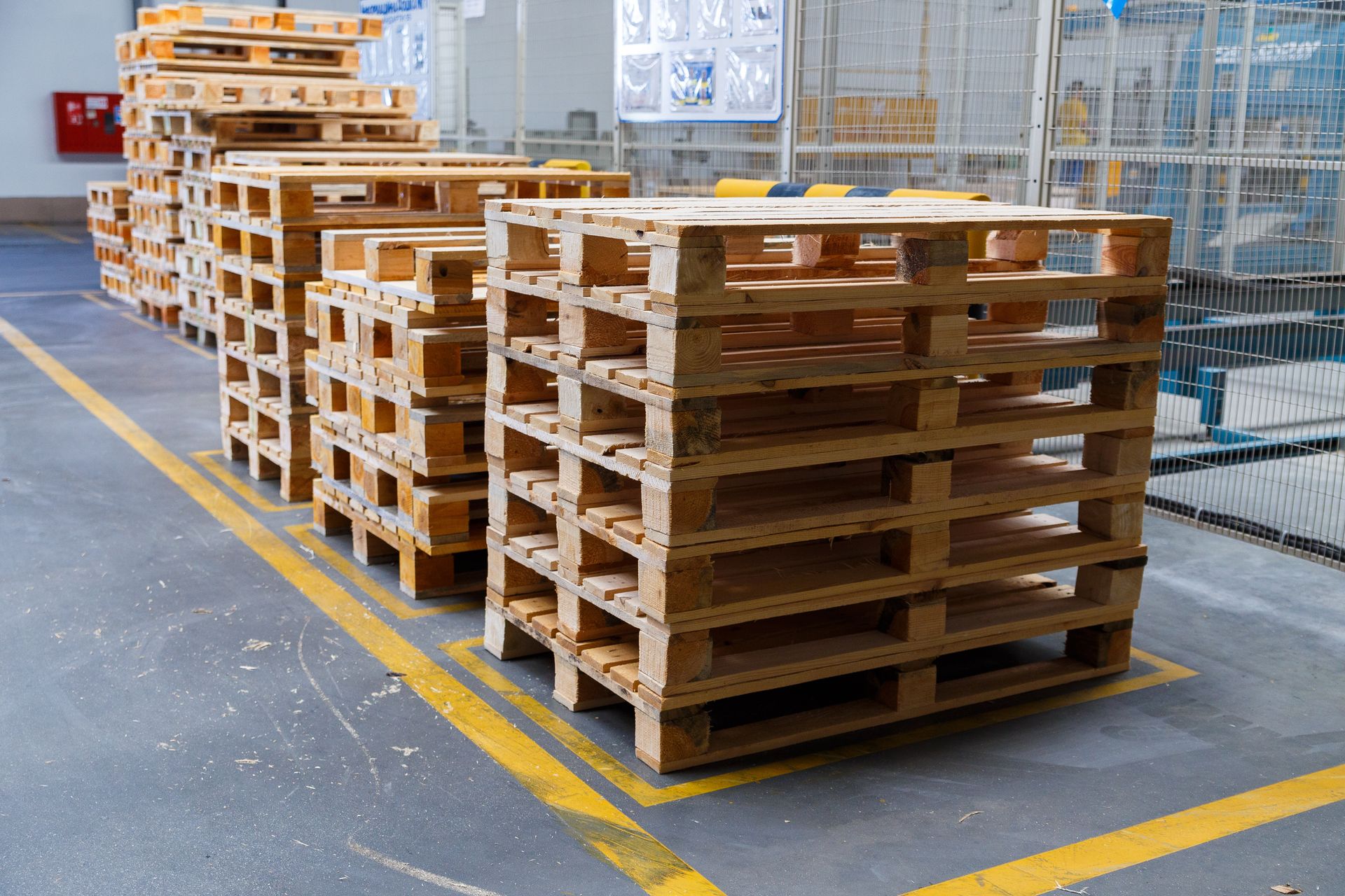 Several stacks of wooden shipping pallets sit on a grey floor marked with yellow safety lines in a warehouse.