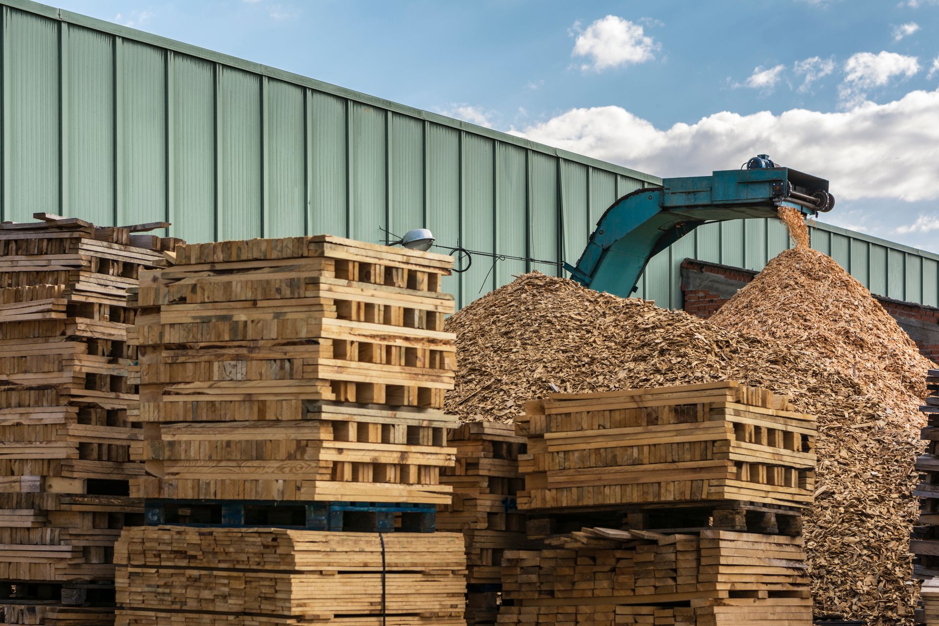 Stacks of wooden shipping pallets sit beside a large pile of wood chips being processed by a machine outside a warehouse.
