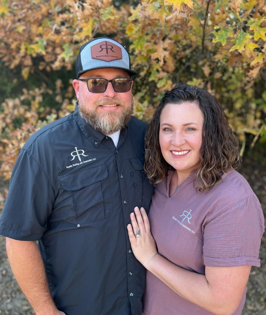 Couple smiling, man wearing a hat and sunglasses, woman with hand on his chest, standing outside.