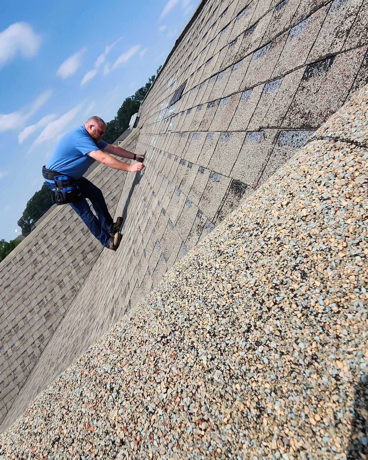 Man on a steep, shingle roof, wearing a safety harness, securing something. Blue shirt, sunny sky.