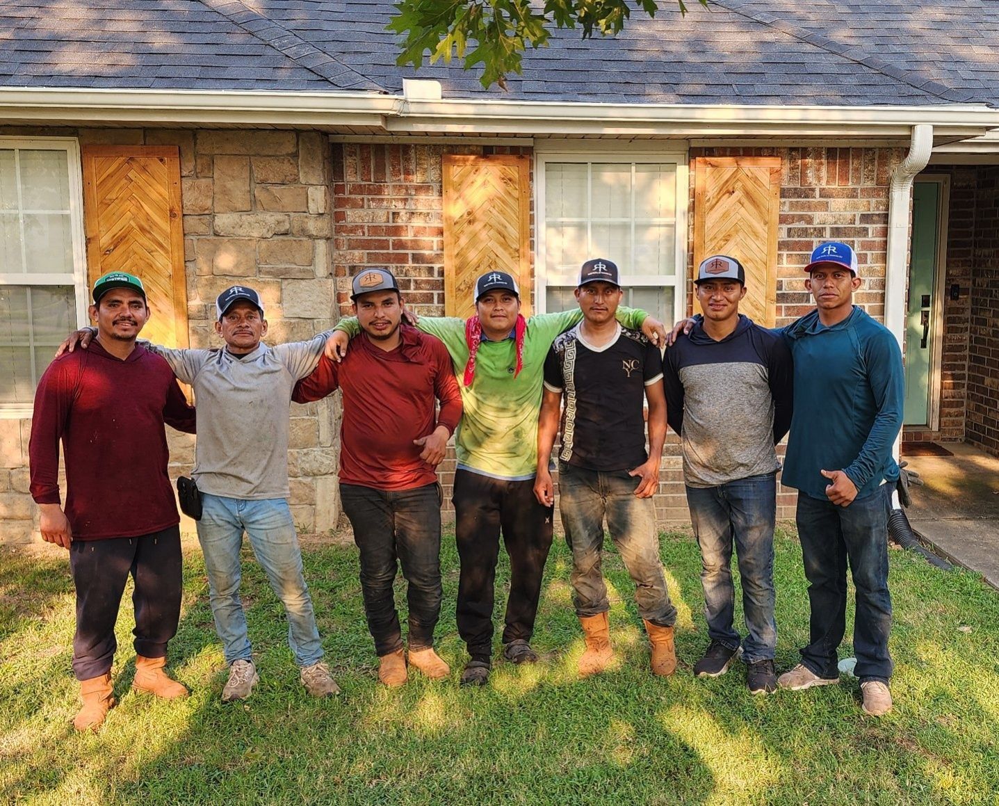Group of seven men, arms around each other, standing in front of a house, wearing hats and work boots.