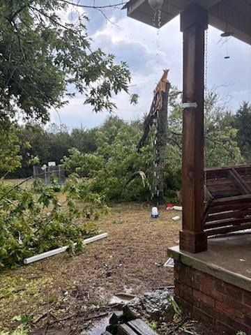 A damaged tree stands in a yard next to a porch with debris, suggesting storm damage.