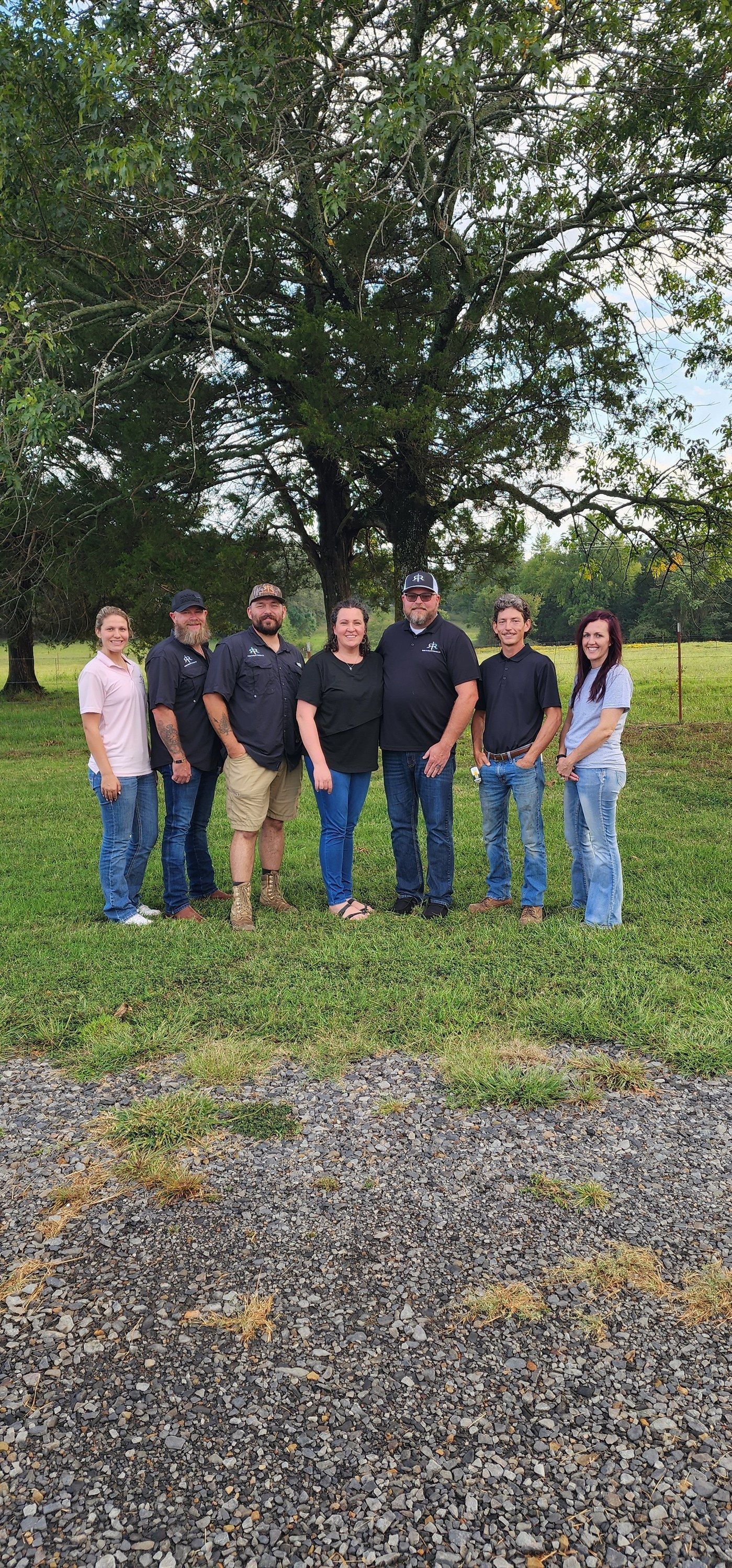 Group of people standing in a field.  Large tree in the background. Green grass and rocks in the foreground.