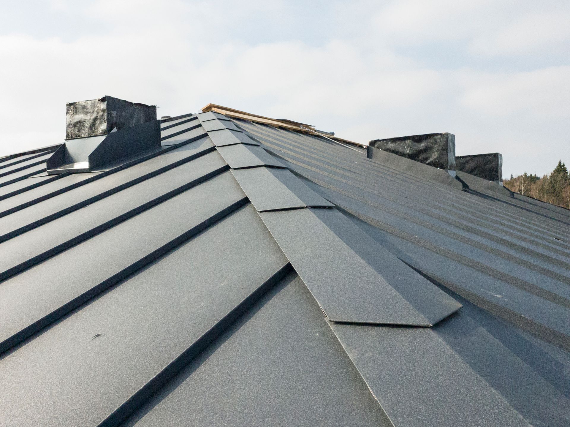 Close-up view of a dark metal roof with rectangular tiles and chimneys against a cloudy sky.