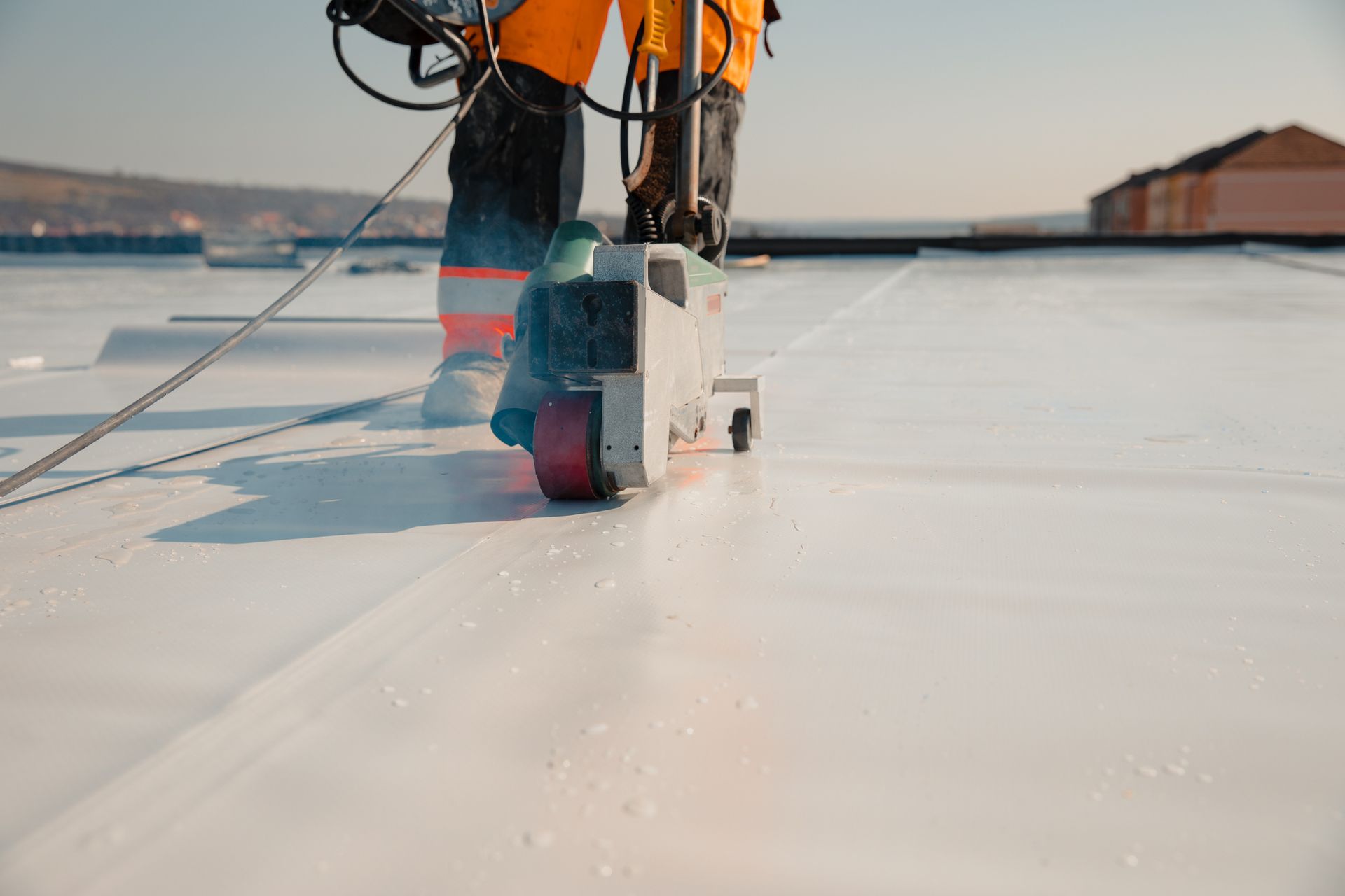 Worker on a white rooftop welds seams with a machine, wearing safety gear.