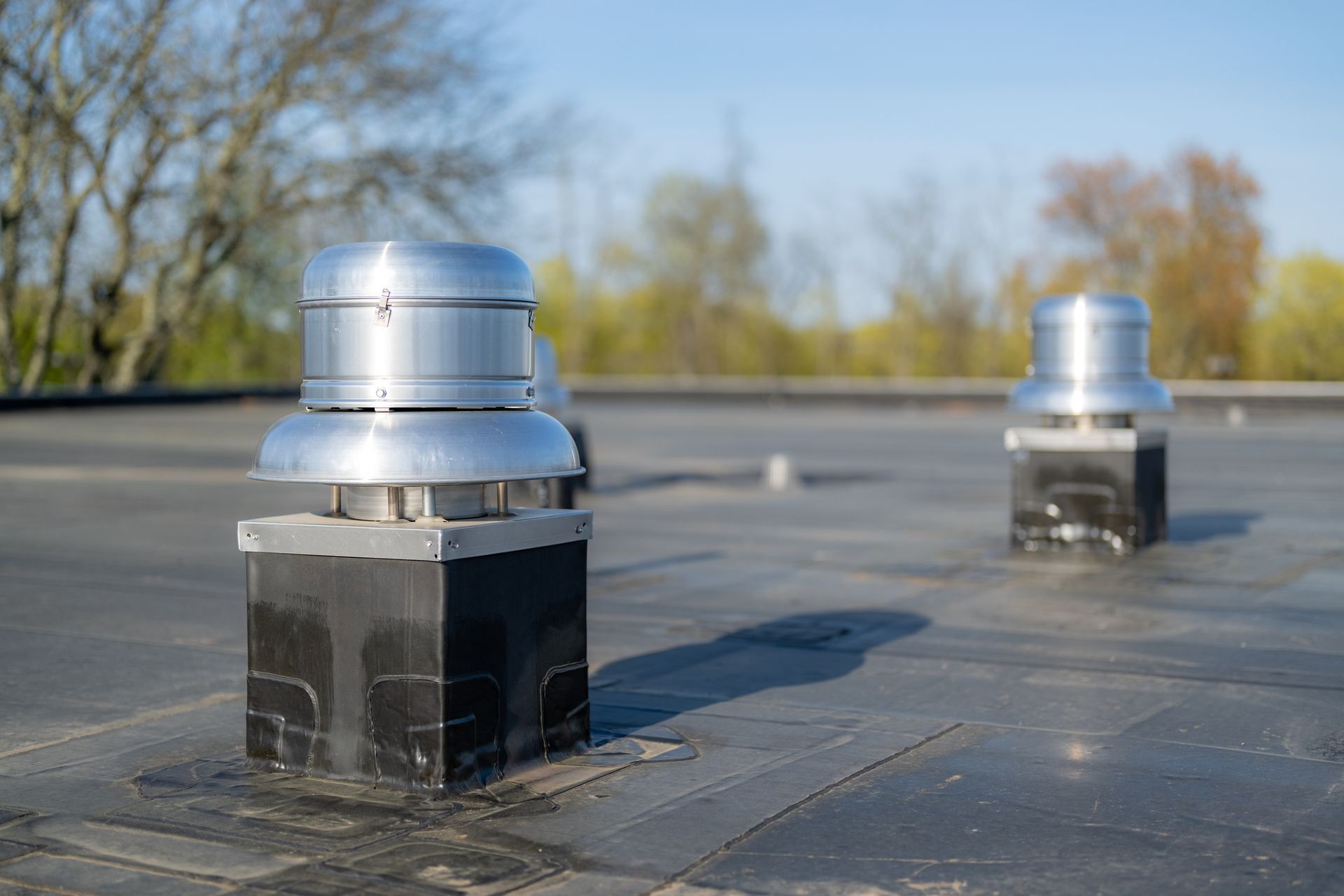 Two metal roof vents on a flat, black roof. Trees and blue sky in background.