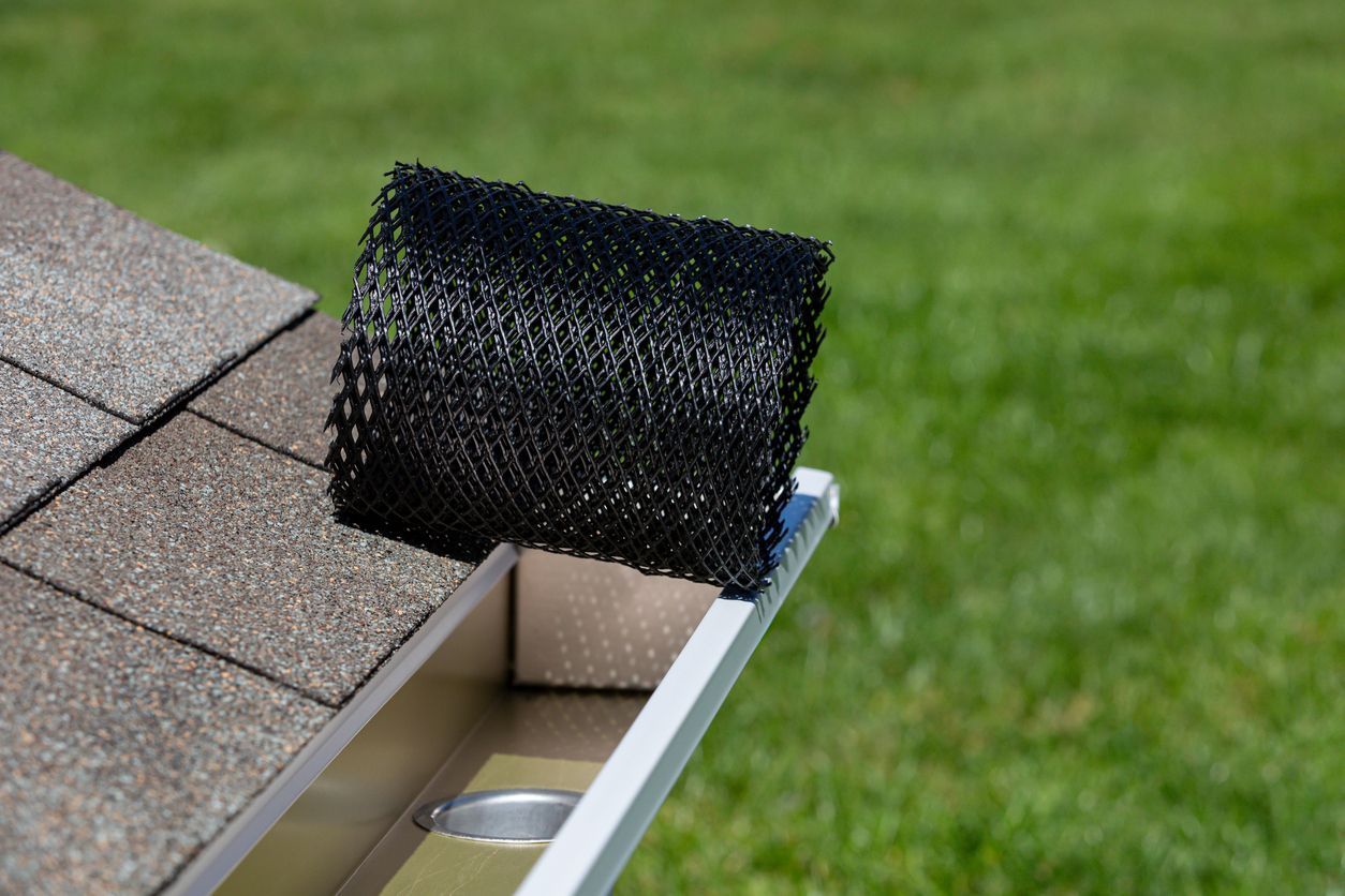 Black gutter guard roll on a roof edge, next to a gutter. Green lawn in the background.