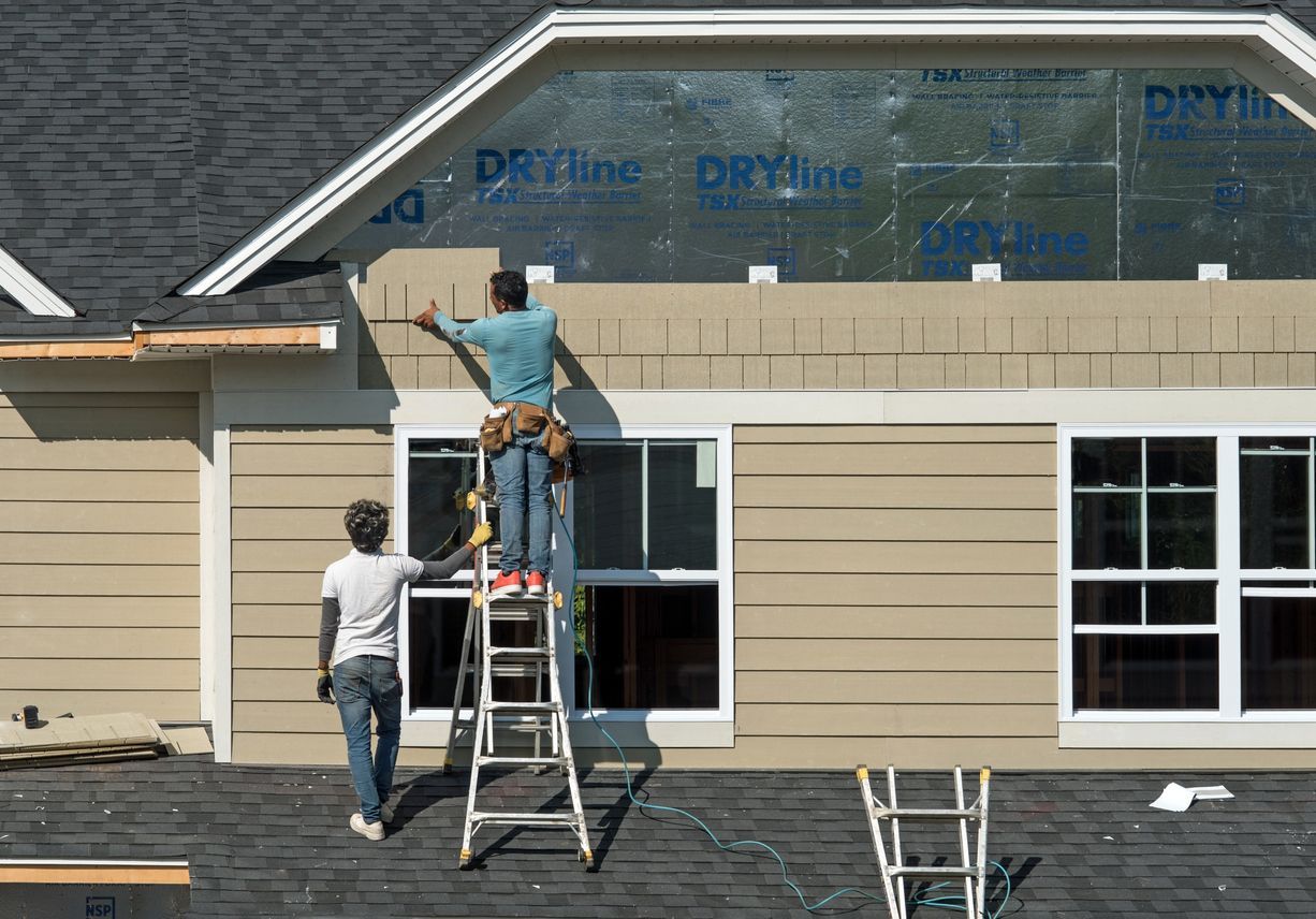 Two workers installing siding on a house exterior; one on a ladder, the other handing a board.