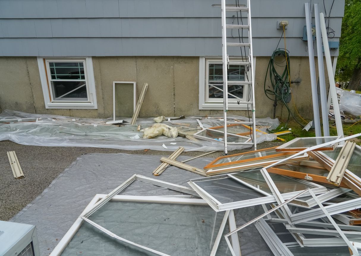 Exterior scene with window replacement in progress; old windows piled on a patio, ladder against the house.