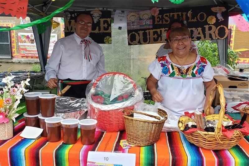 A man and a woman are standing behind a table with a sign that says queso taro