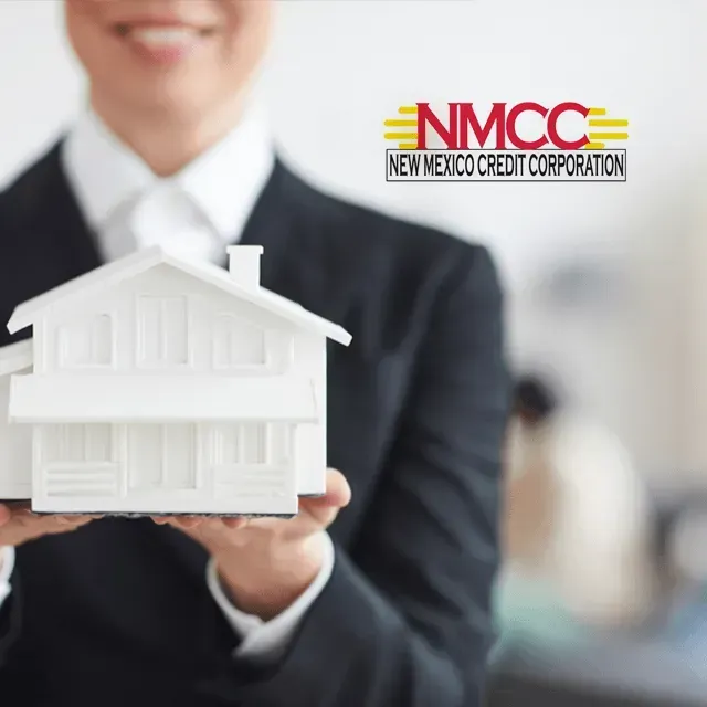 A woman in a suit is holding a model house with the nmcc new mexico credit corporation logo in the background