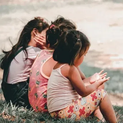 Three little girls are sitting in the grass looking at the ocean.