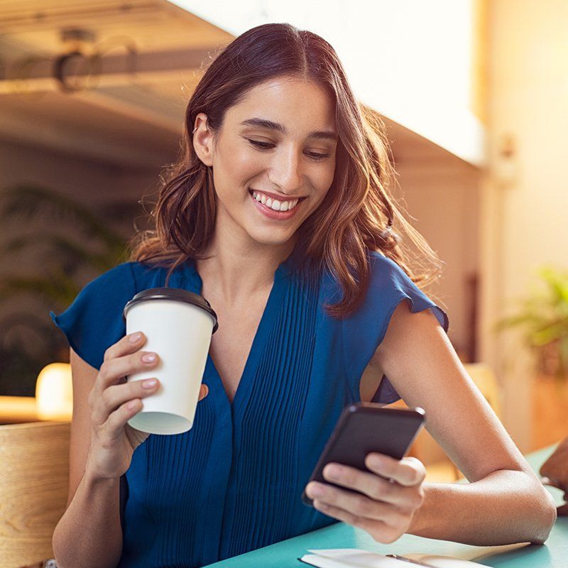 A woman is sitting at a table holding a cup of coffee and looking at her phone.