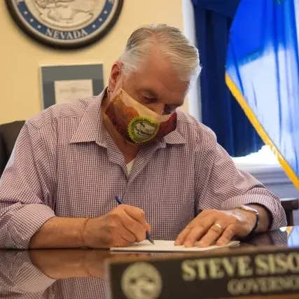 A man wearing a mask sits at a desk with a name plate that says steve siso