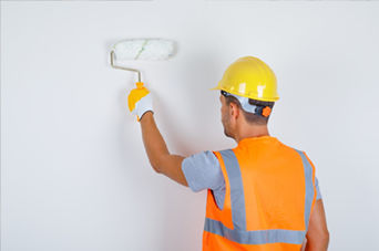 Person in hard hat and orange vest painting white wall with a paint roller.