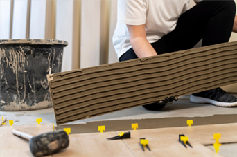 Person installing floor tiles, applying adhesive to tile, using levelers and a mallet indoors.