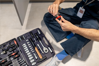 Person seated with toolbox, holding red and black tool; wearing blue pants and socks.
