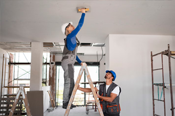 Two construction workers sanding a ceiling. One on a ladder, the other supporting it. Inside a building under construction.