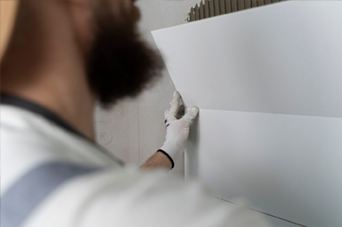 Person in orange safety vest and yellow hard hat using a paint roller on a white wall.