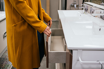 Person in mustard coat opens bathroom vanity drawer in a showroom.