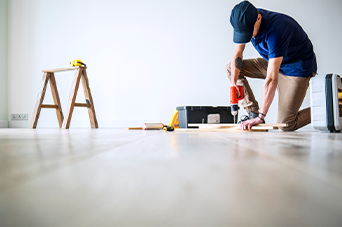 Man in a cap, kneeling and using a drill on a wooden floor in a room. A toolbox and stepladder are nearby.