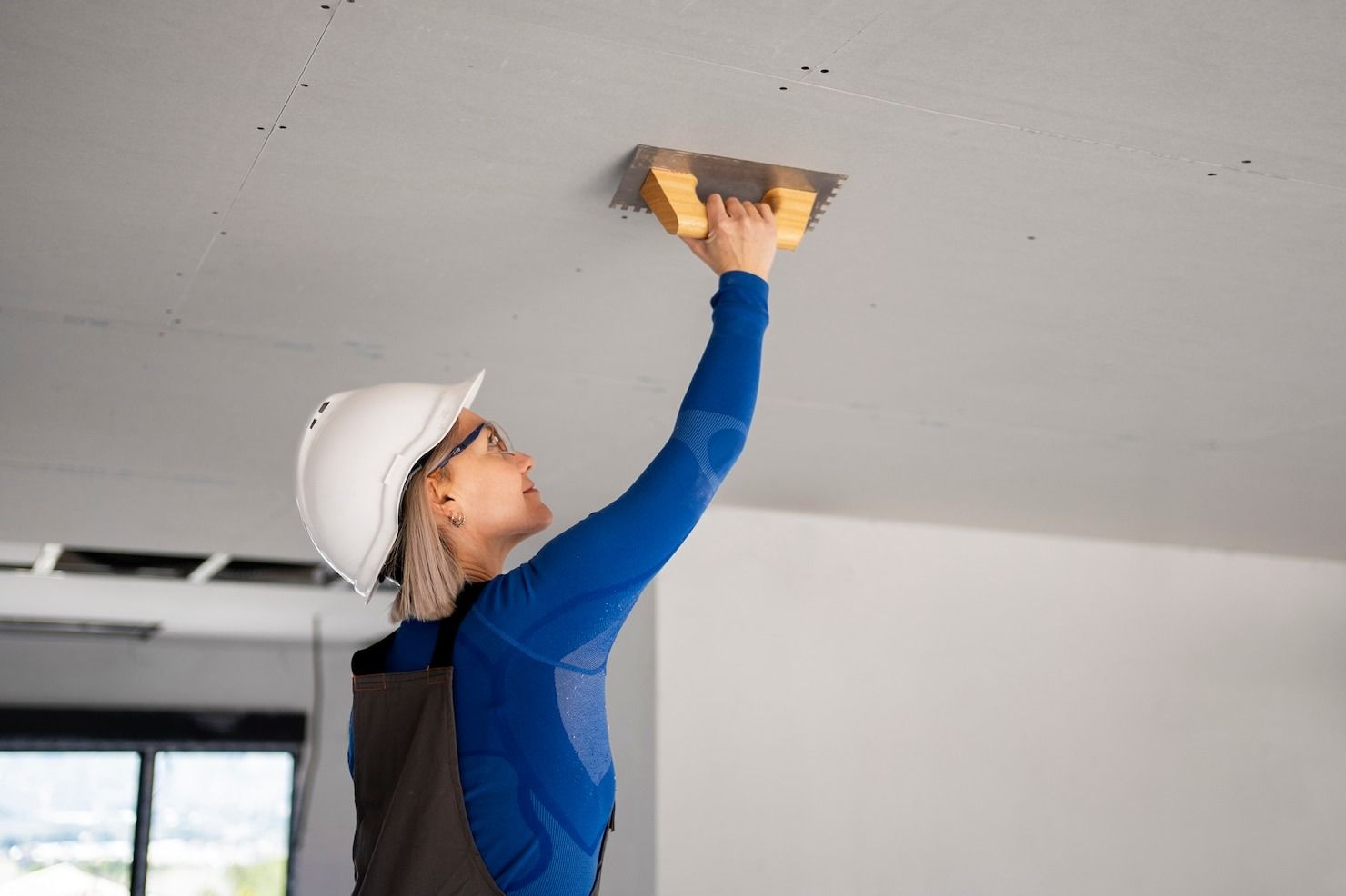 Person installing flooring, holding a plank covered with adhesive. Tools and bucket nearby.