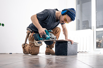 Person kneeling, sawing wood with a power saw. Wearing safety glasses and ear protection in a room.