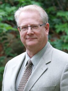 A man in a suit and tie is standing in front of trees.