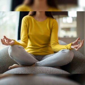 A woman in a yellow shirt is sitting in a lotus position on a couch.