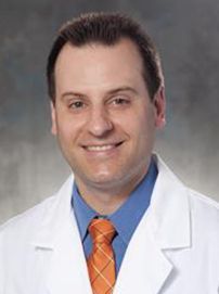 A man in a white coat and orange tie is smiling for the camera.