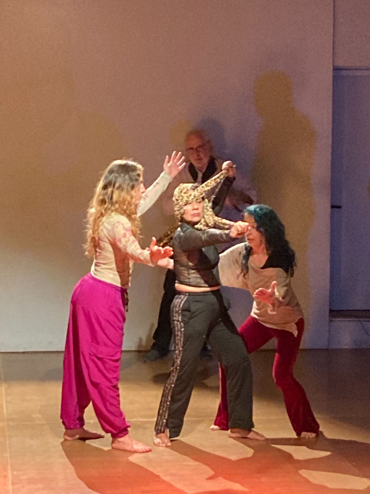 Tres bailarines vestidos con ropa de baile informal se mueven juntos en un estudio, con una persona parcialmente visible al fondo.