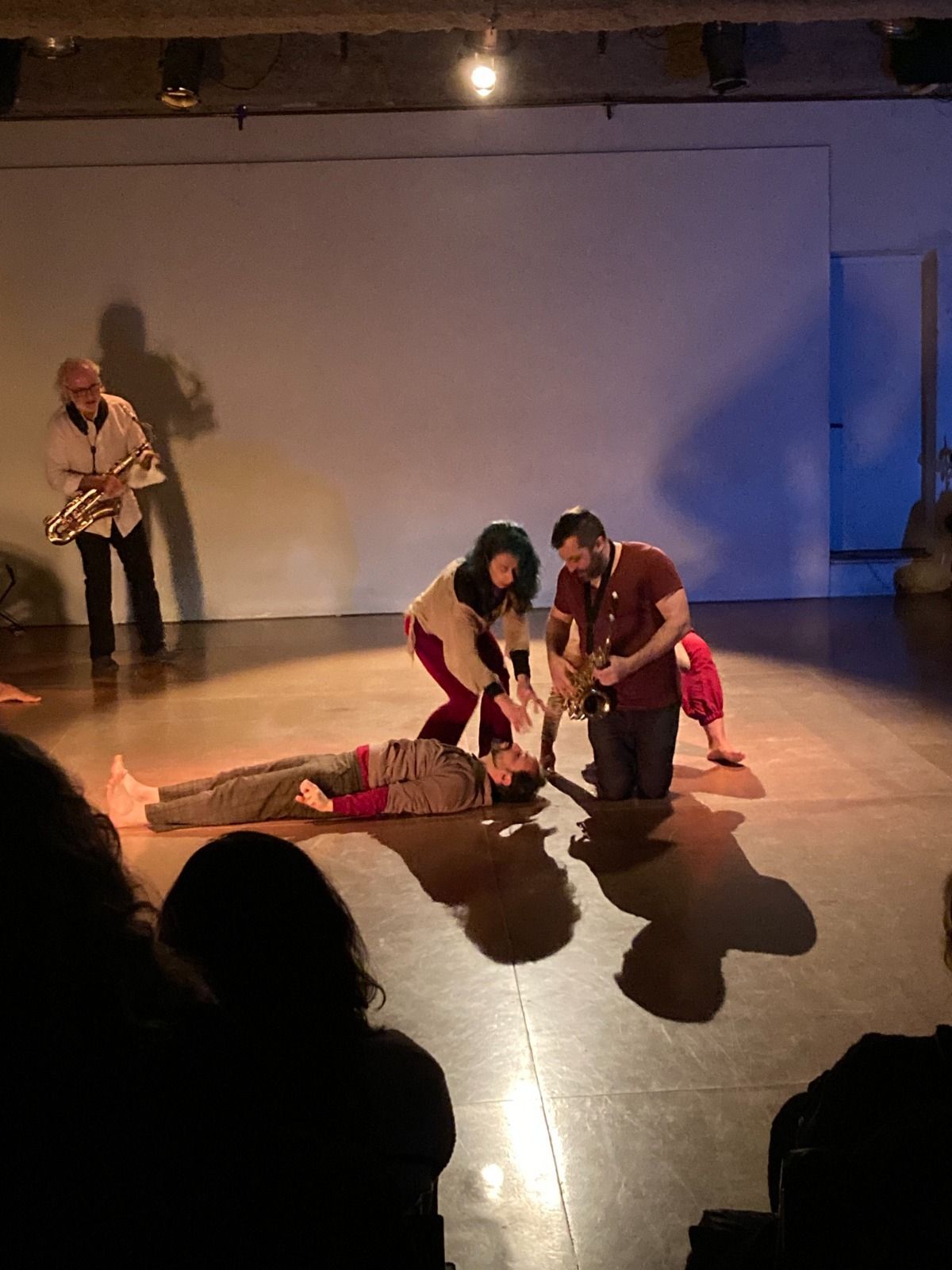 Cuatro artistas en un escenario: uno toca el saxofón, otro está tumbado en el suelo y dos se arrodillan junto a ellos bajo un foco.