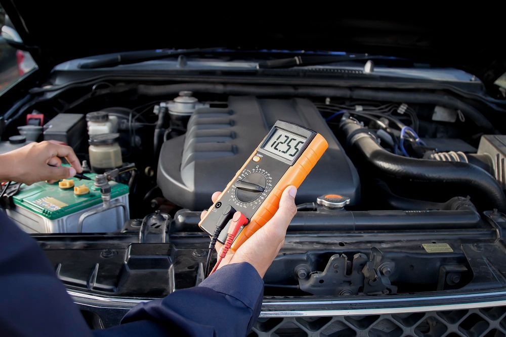 Person using a multimeter to test a car battery in an open engine bay, showing a voltage reading of 12.55 volts.