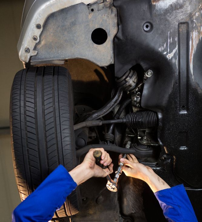 Mechanic working on a car's undercarriage; blue uniform, hands using tools near tire and steering components.