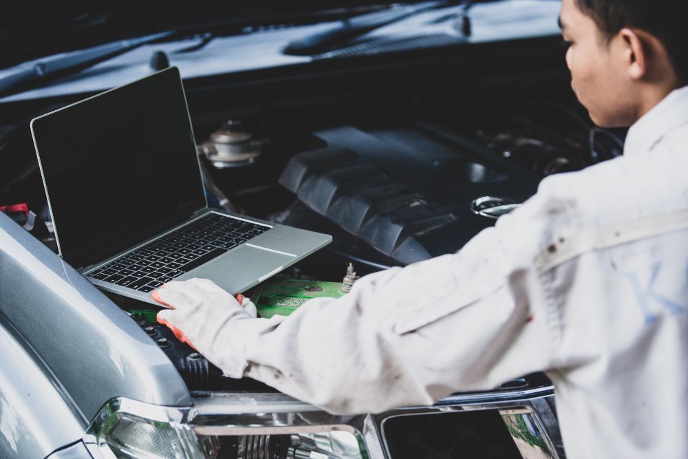 Mechanic using a laptop to diagnose a car engine with the hood open.