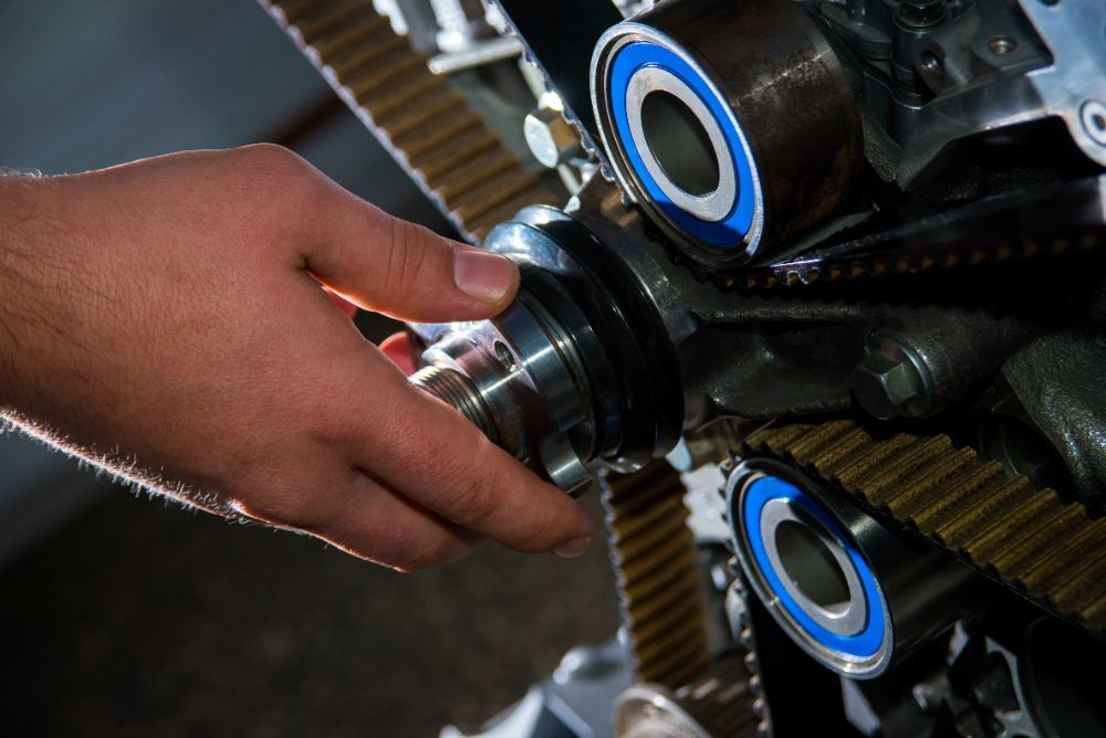 Hand adjusting a silver metal piece in an engine, near belts and blue-rimmed bearings.