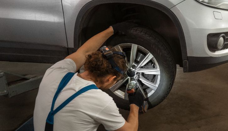 Mechanic uses a wrench on a car tire, working beneath a silver vehicle in a garage.