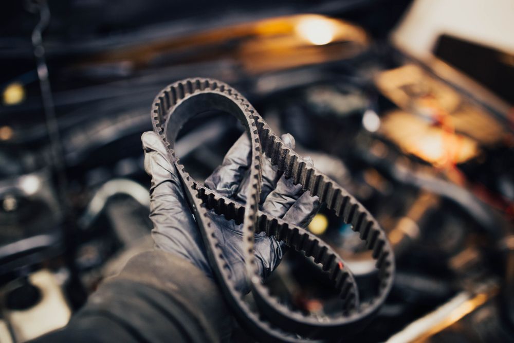 A gloved hand holds a worn, toothed timing belt against the backdrop of an open car engine bay.