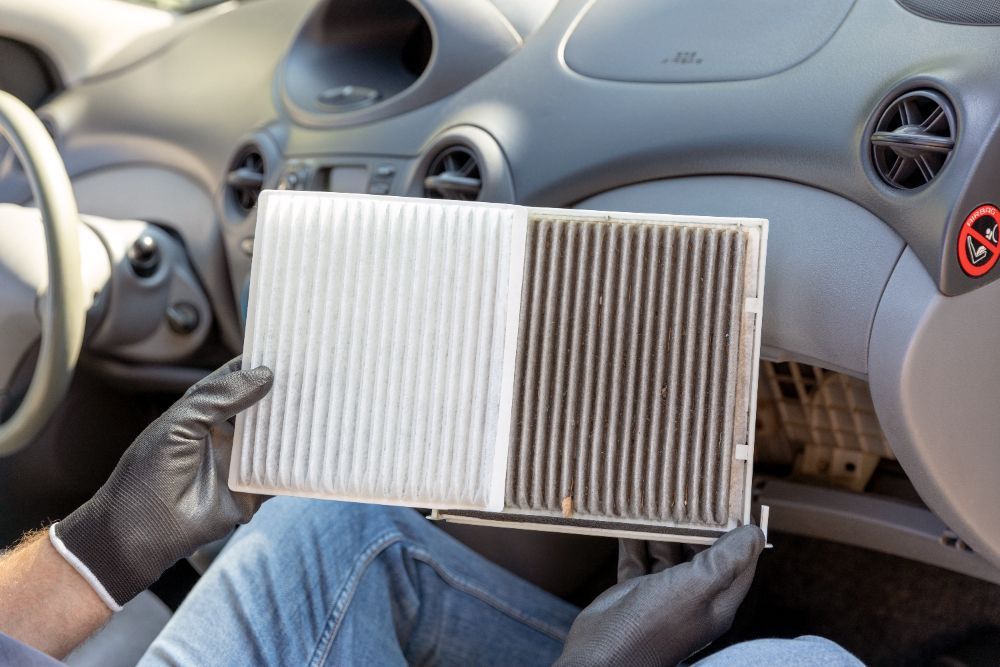 Hands holding a clean and dirty cabin air filter in a car dashboard.