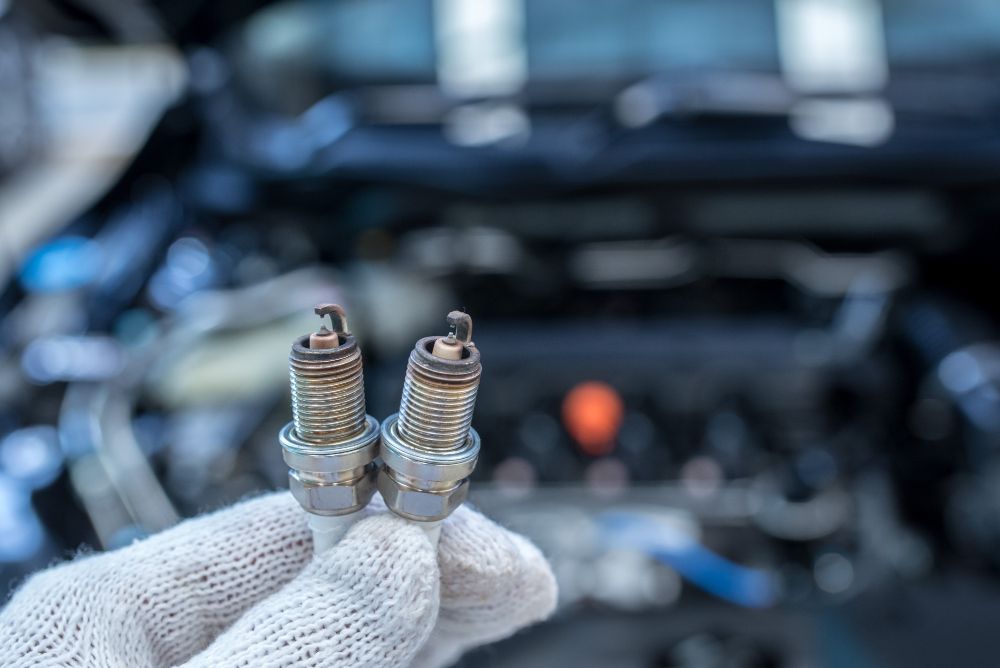 Gloved hand holding two used spark plugs in front of a car engine bay.