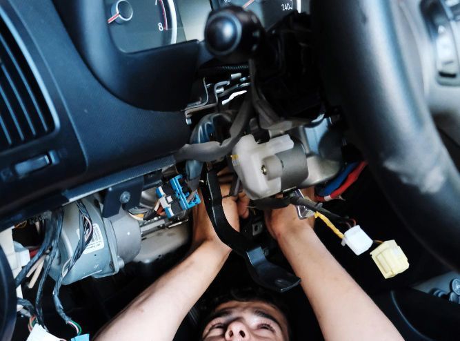 Person working on car's steering column, hands visible, wires and components in view.
