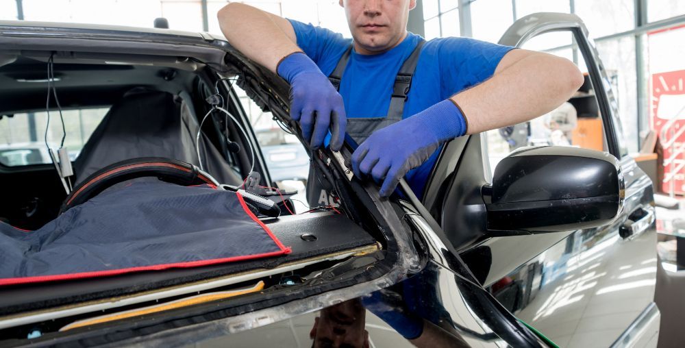 A mechanic in blue gloves and shirt replacing a car windshield in a repair shop.