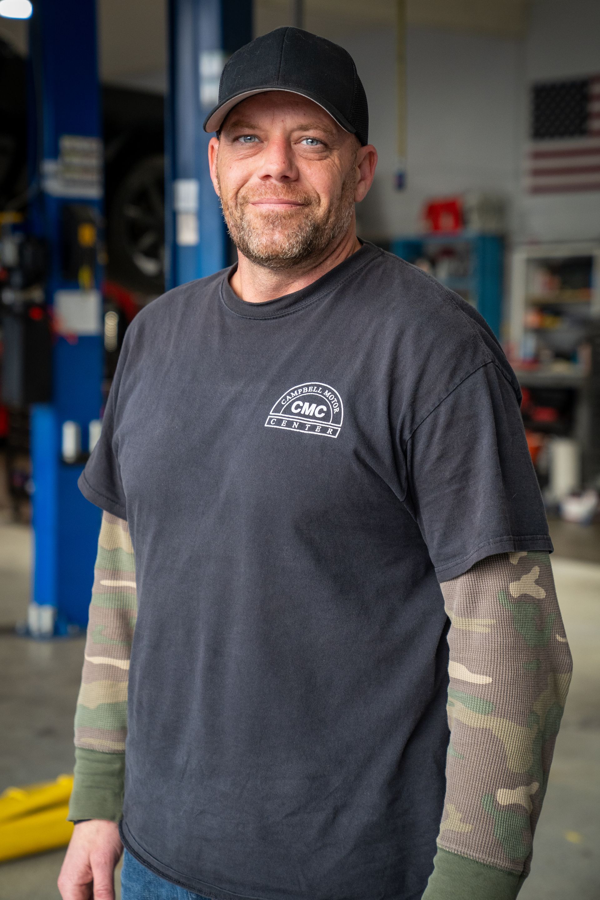 Man in a garage wearing a black t-shirt and camo sleeves, looking at the camera.