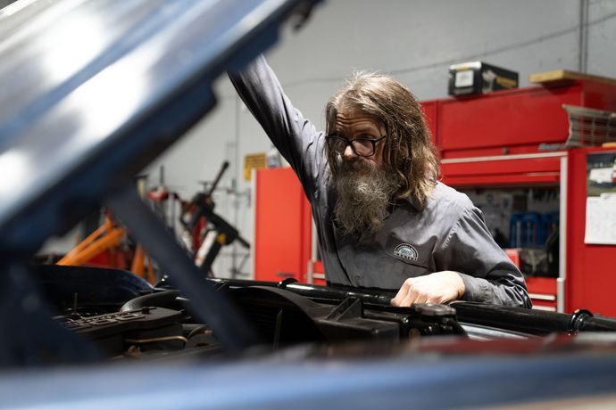 Mechanic with long hair and beard working under the hood of a car in a garage.